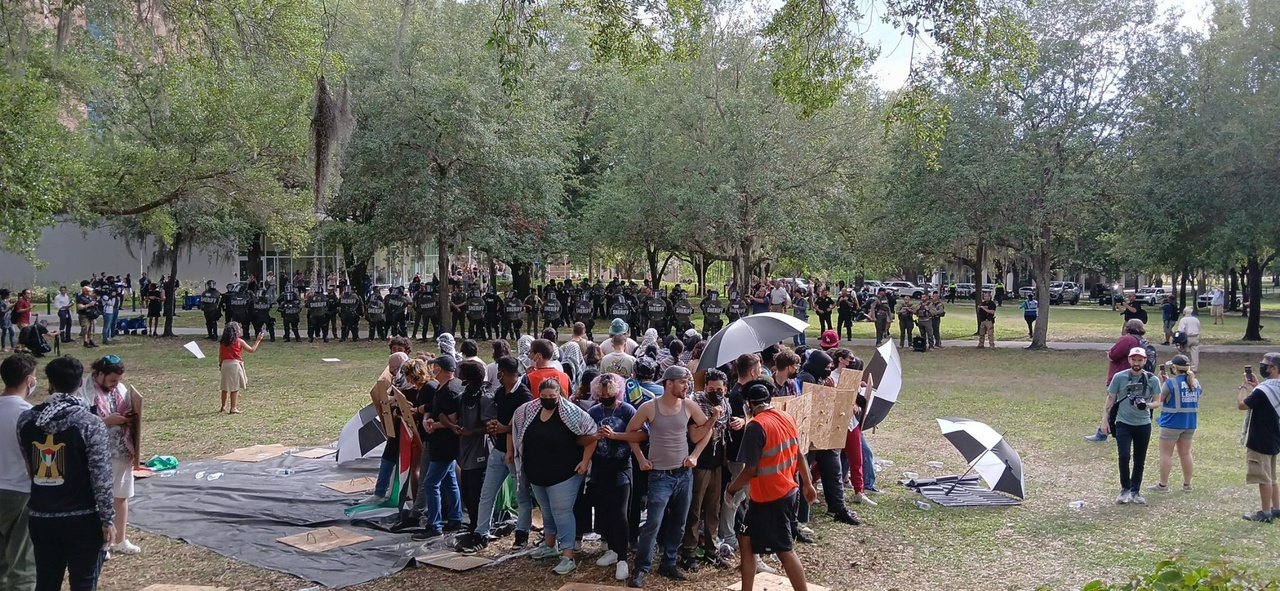 Group of protesters stand in a circle facing outward with their elbows interlocked. In the background several lines of police officer with riot shields are visible.