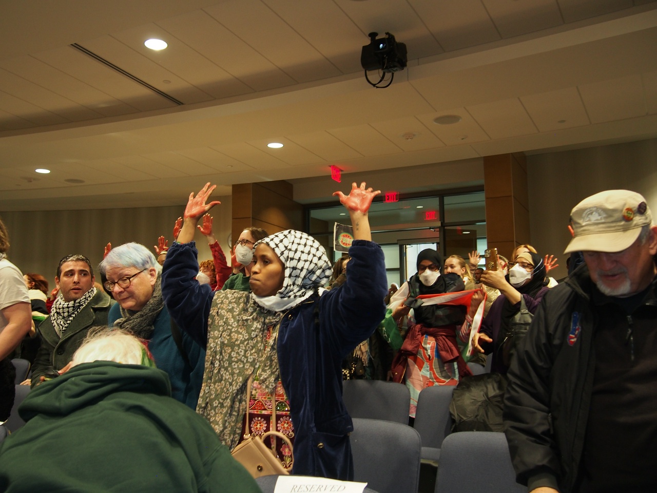 Group of protesters wearing keffiyehs and holding Palestinian flags raise their hands to disrupt a meeting.