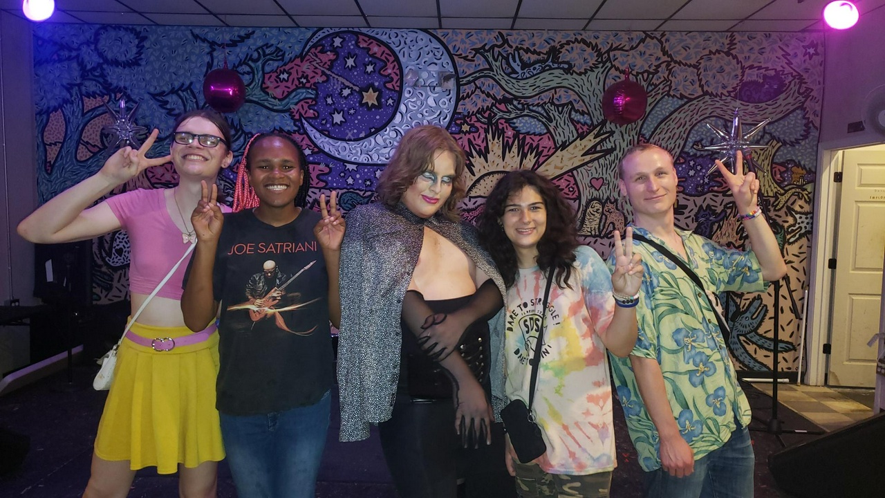 Five young queer and trans people of hold up peace signs and pose smiling.