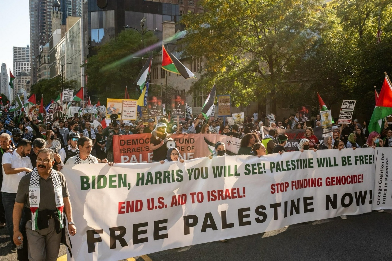 Large crowd gathers behind a banner that reads “BIDEN, HARRIS YOU WILL SEE! PALESTINE WILL BE FREE! END US AID TO ISRAEL! STOP FUNDING GENOCIDE! FREE PALESTINE NOW!”