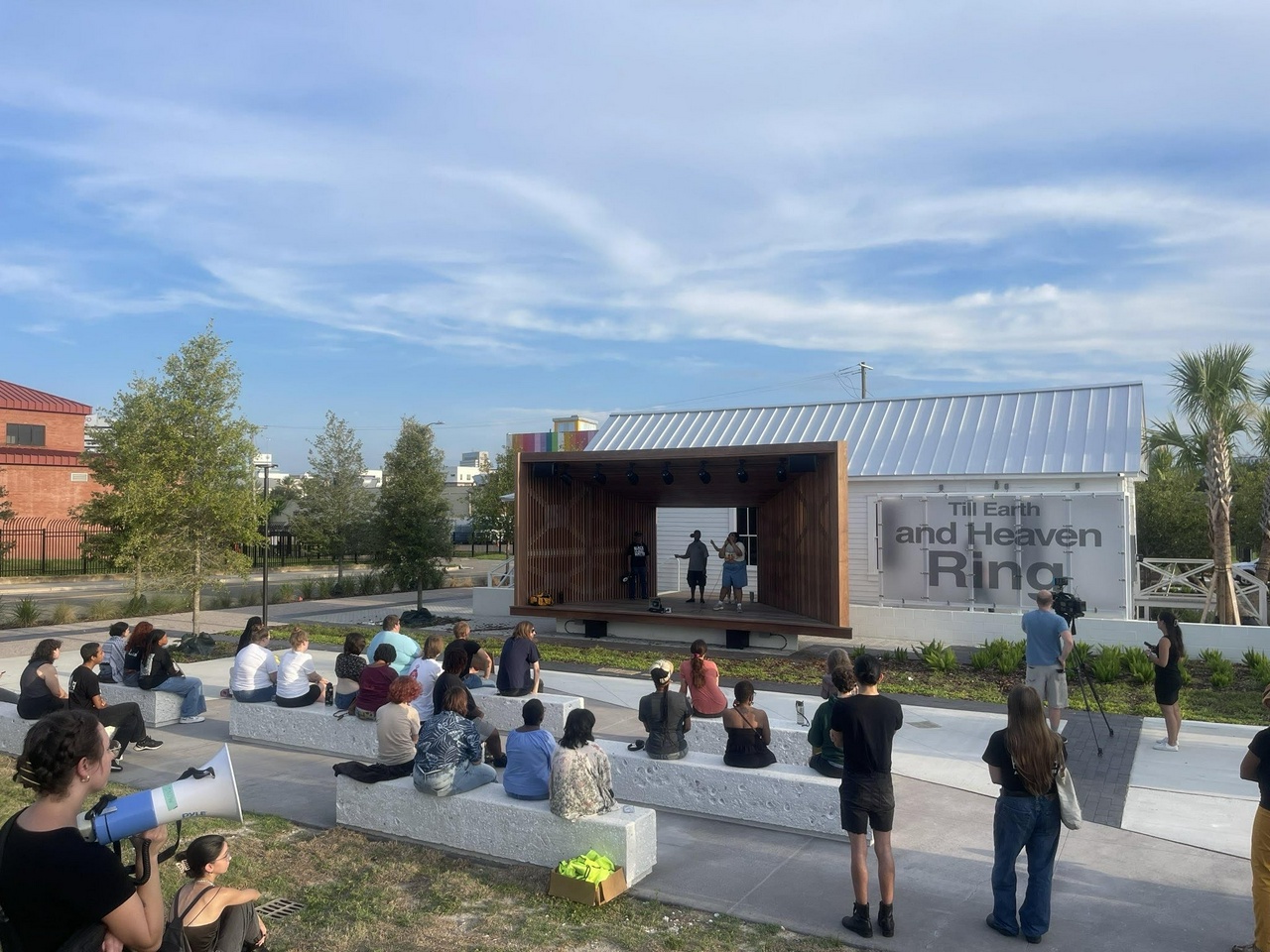 About 20 people sit in an outdoor amphitheater while two people speak on microphones on a stage.
