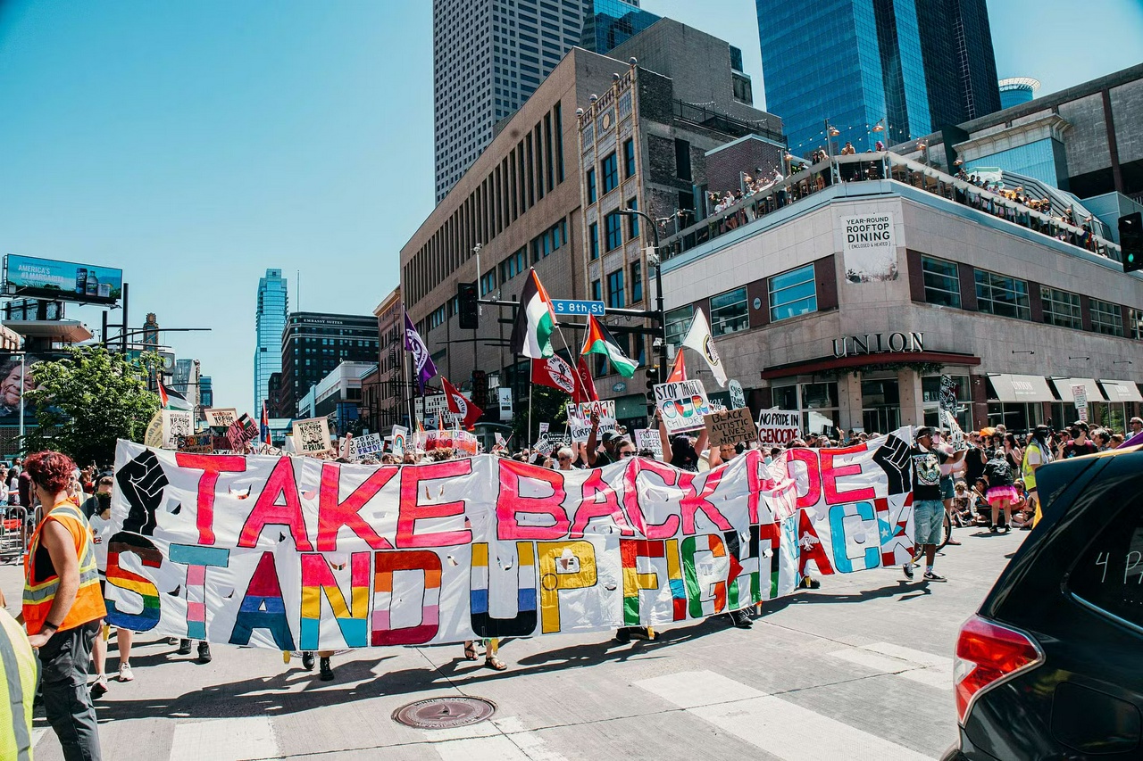 Protestors march through the street with an audience watching on the sidewalks. The march carries a large banner that reads "Take Back Pride. Stand Up, Fight Back".