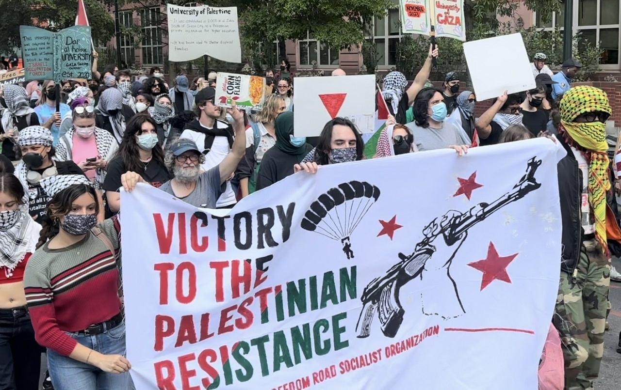 Crowd marches behind banner that reads “Victory to the Palestinian Resistance”.