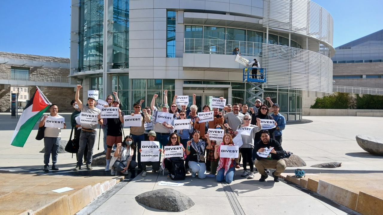 Demanding divestment from apartheid Israel in San Jose, California. | Fight Back! News staff A group of people, some standing and some kneeling, in front of a building. They are holding signs that read, 'DIVEST'. One person holds a Palestinean flag.