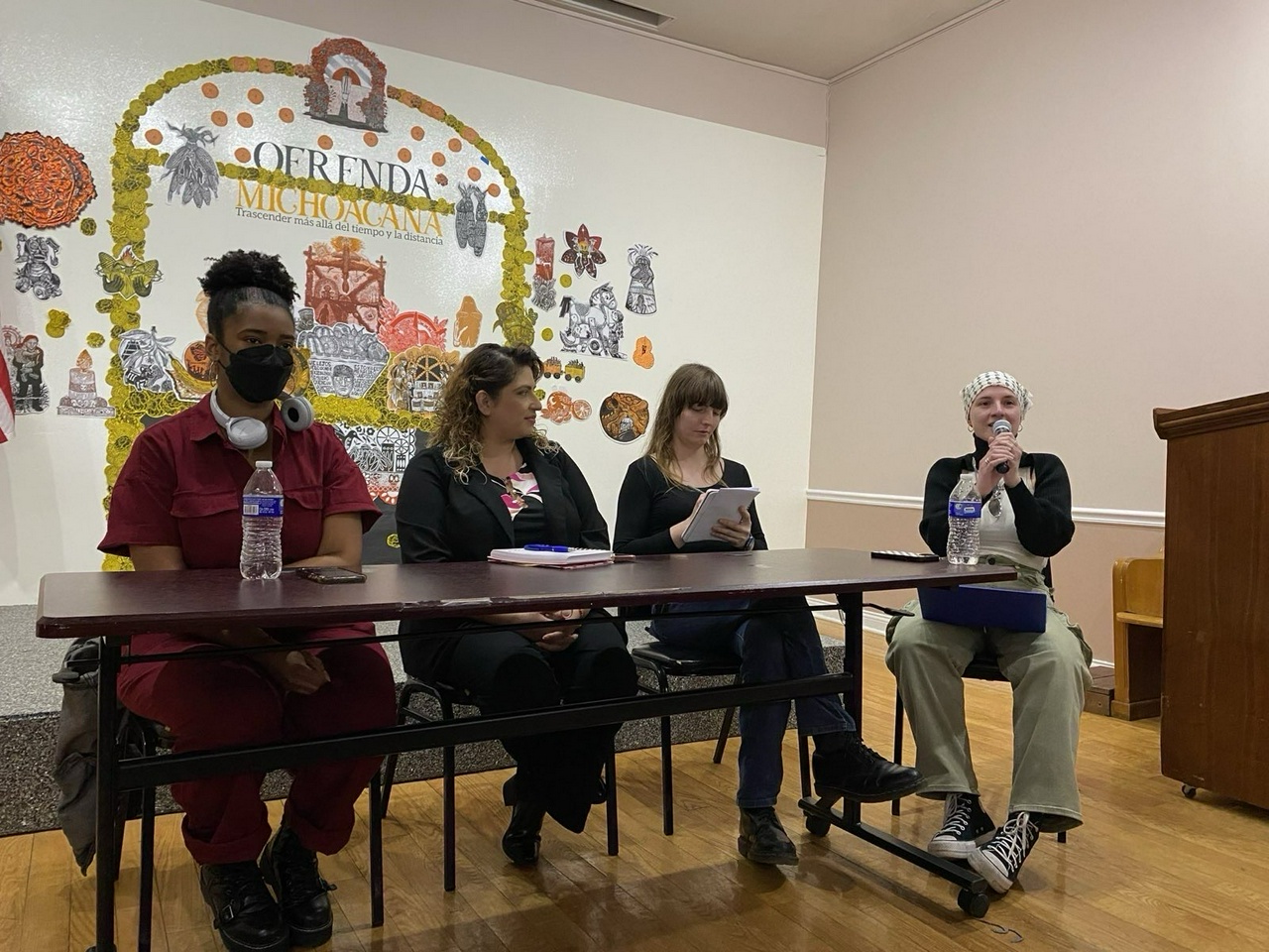 Panelists sit at a table in front of a mural of an Ofrenda. One panelist wearing a keffiyeh holds a mic and speaks.