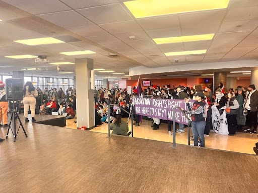 Crowd gathers indoors, a banner is prominent that reads “ABORTION RIGHTS”