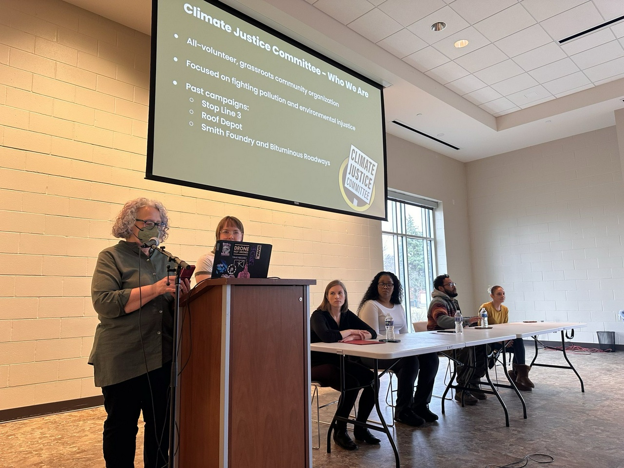 Two people stand behind a podium, speaking into a microphone. Nearby, four people sit at a table. There is a projector screen above reading, "Climate Justice Committee - Who We Are"