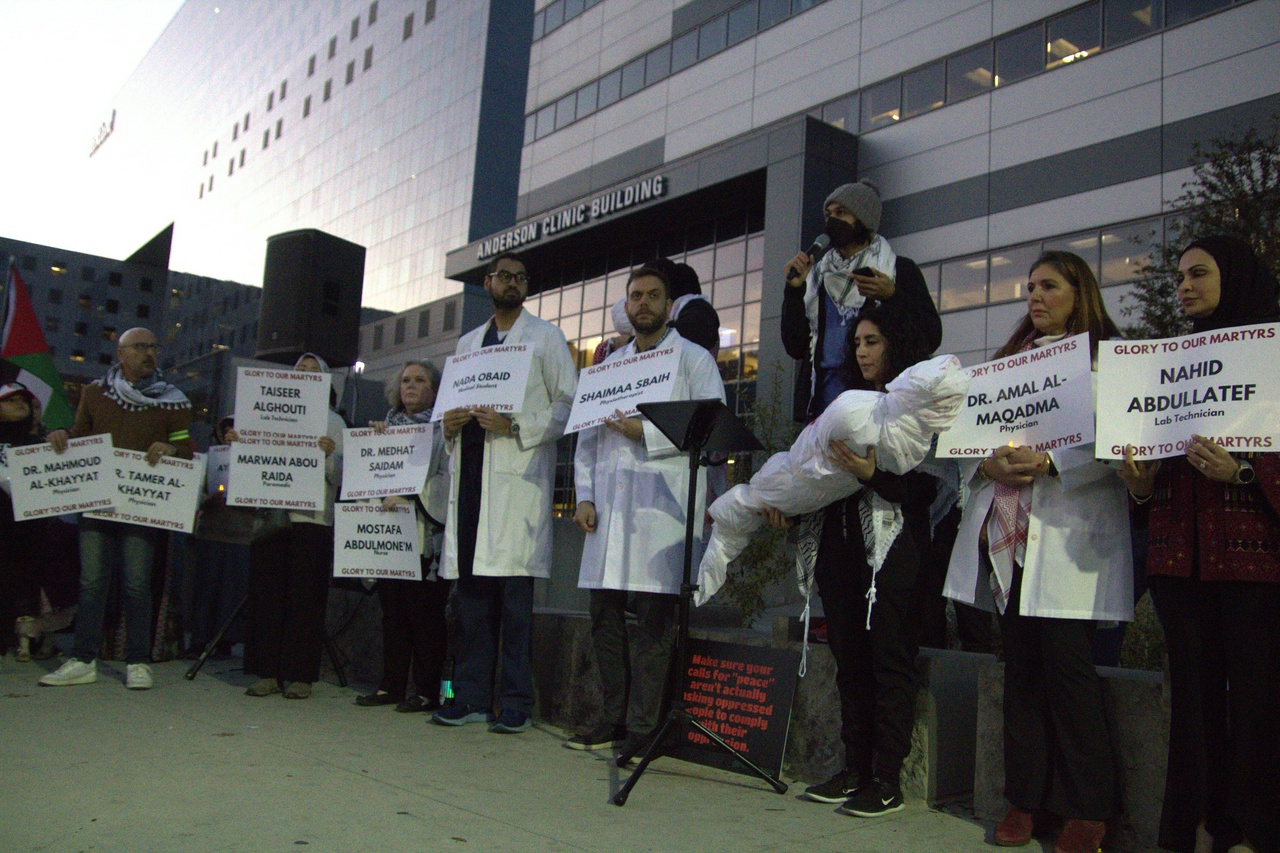 A line of people wearing lab coats hold signs with the names of medical professionals martyred in Gaza. One person holds a human-sized shroud wrapped in white cloth.