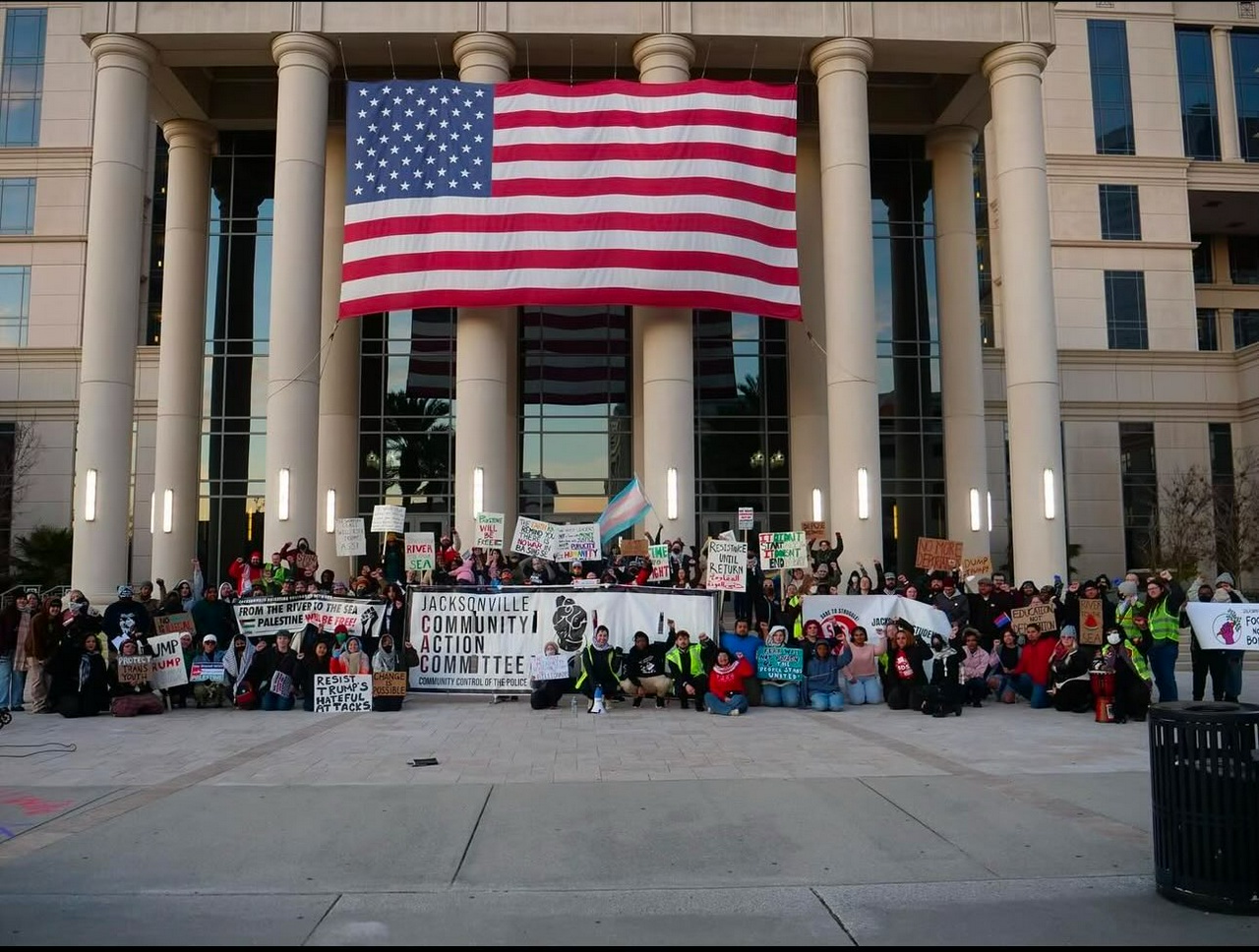 Crowd gathers on courthouse steps beneath massive U.S. flag. 