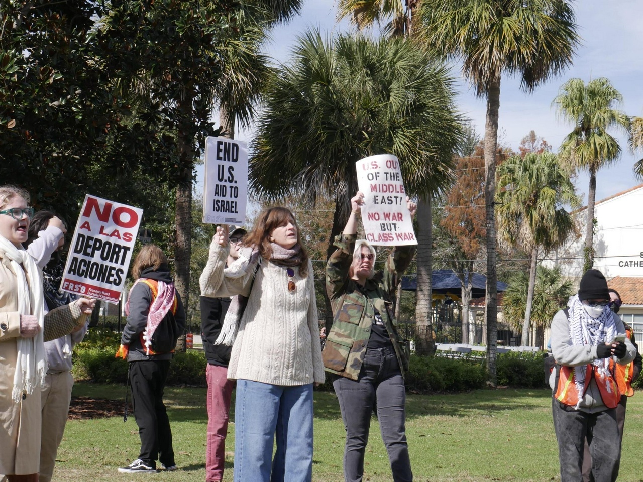 Protesters hold signs on a green. Their signs have slogans like “US out of the Middle East” and “No a las deportaciones”.