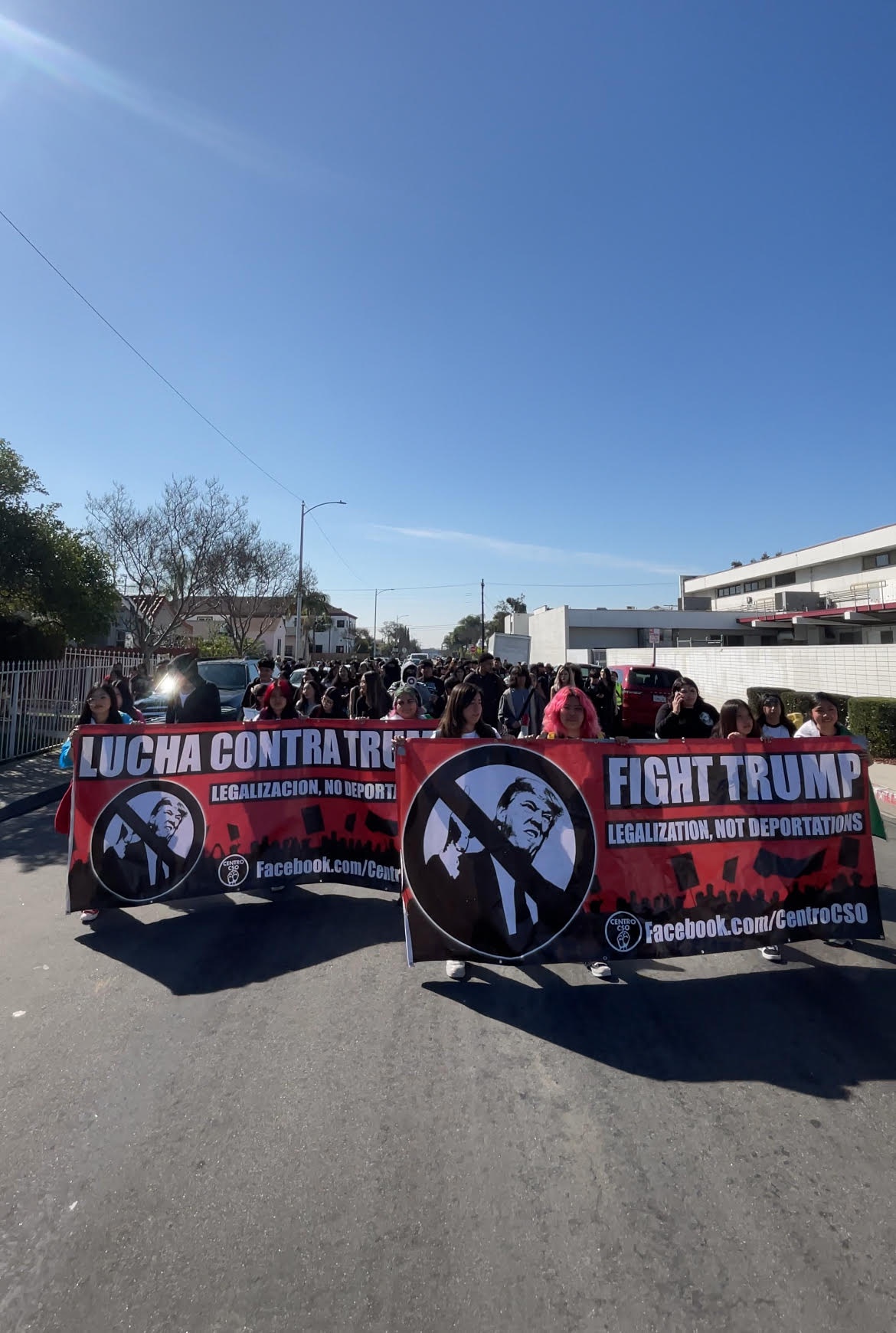 Students march in street. Students at front hold two banners, one in saying “LUCHA CONTRA TRUMP, LEGALIZACION, NO DEPORTACIONES” and the other reading “FIGHT TRUMP, LEGALIZATION, NOT DEPORTATIONS”.