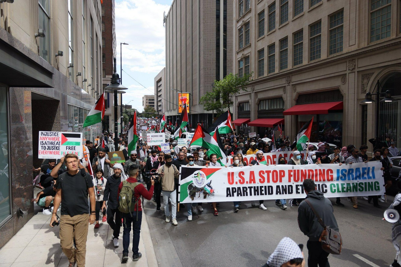 Hundreds of people are visible holding Palestinian flags march down a city street behind a banner that reads: "USA Stop Funding Israeli Apartheid and Ethnic Cleansing" 