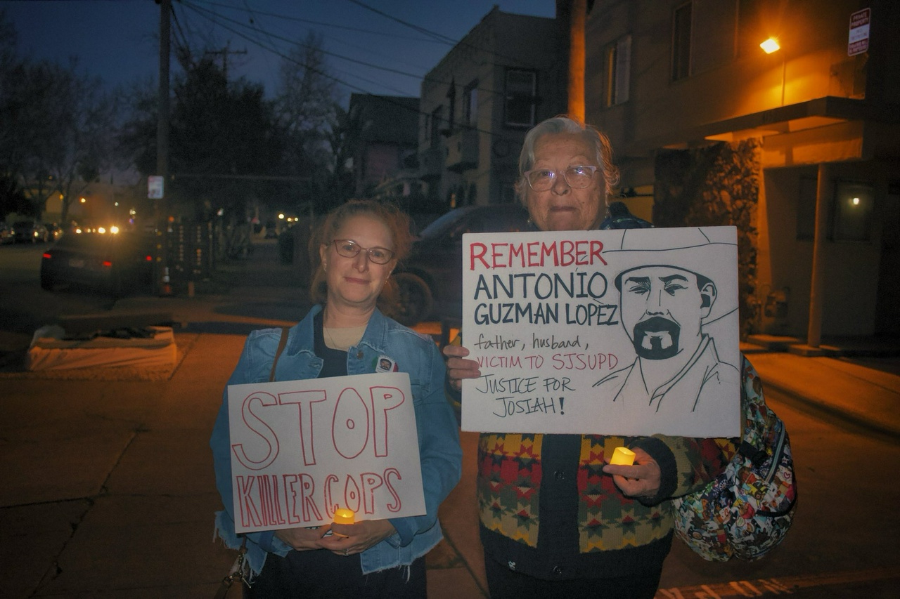 Two women hold signs and candles on a sidewalk at night. One sign says “Stop Killer Cops” and the other has a drawing of Antonio Guzman and says “Remember Antonio Guzman Lopez, father, husband, victim to SJSUPD. Justice for Josiah!”
