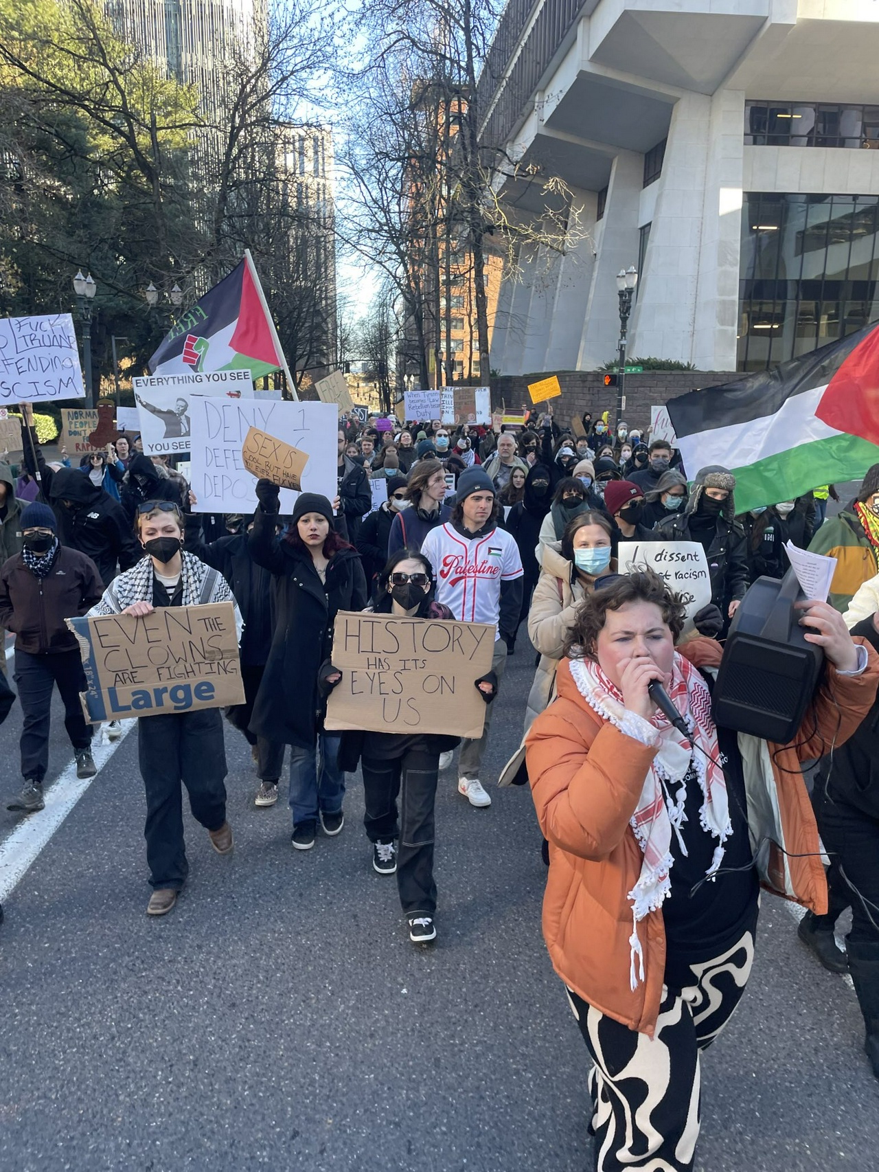 Large group of people marches down the street carrying Palestinian flags and signs with slogans like “History has its eyes on us.”