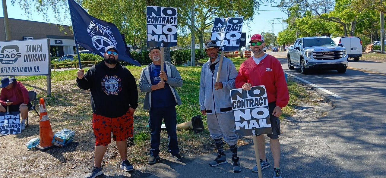 A group of truckers hold signs that say, "No Contract, No Mail" outside of 10 Roads Express workplace.