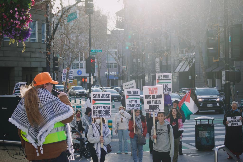 A rgoup of people stands in front of a street. They hold signs and Palestinean flags.