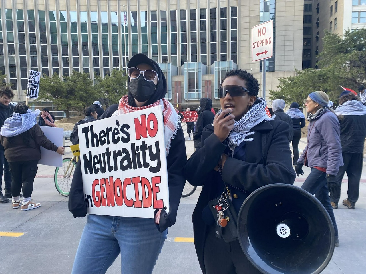 Two people standing in front of a group. One person's sign reads, "There's no neutrality in genocide"