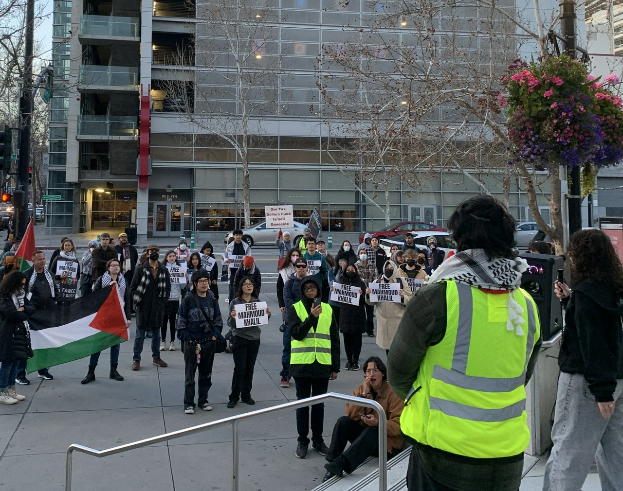 A crowd of people standing on a street corner, some holding Palestinean flags, others hold signs reading "Free Mahmoud Khalil."