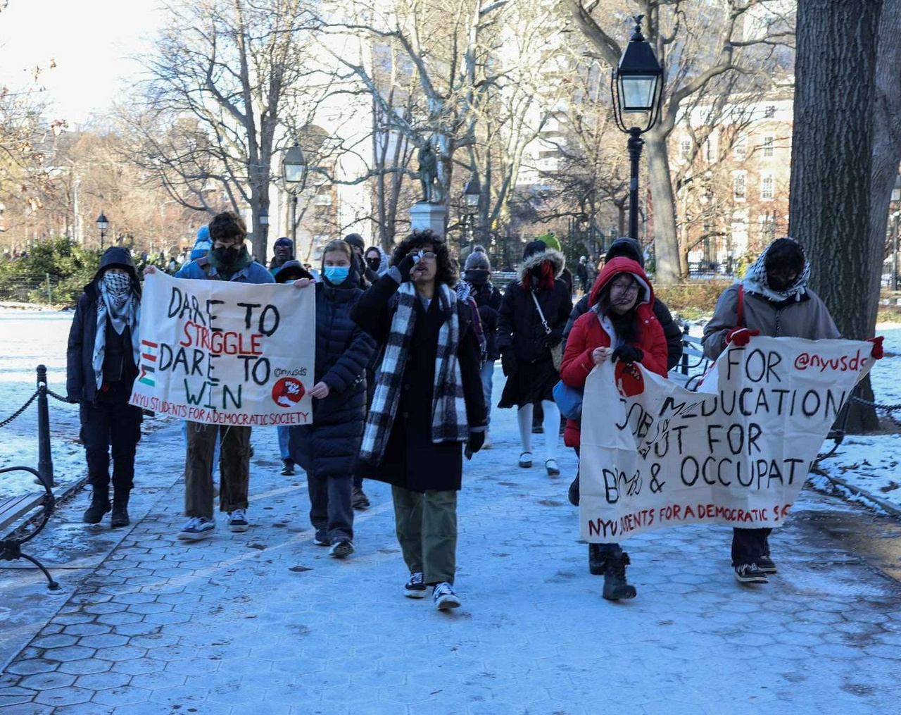Protesters gather in a snowy park with signs reading things like “Dare to Struggle, Dare to Win @NYUSDS”.