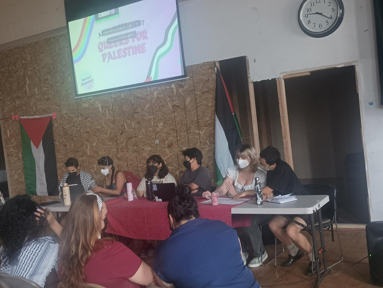 Presenters sit at a table in the front of a seated audience. A slide is projected behind the presenters that says "queers for palestine".