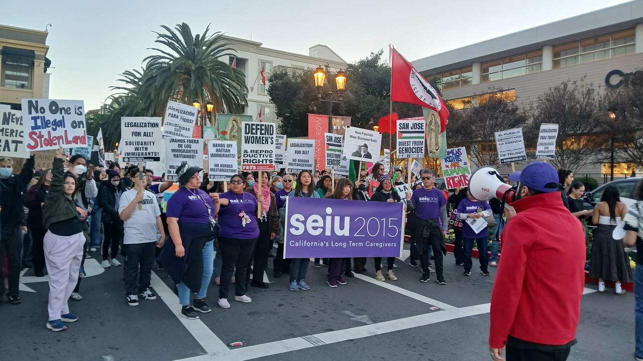 Protesters gather while a lead organizer speaks into a bullhorn facing the crowd. The gathered crowd holds signs with slogans including “No one is illegal on stolen land” and “Legalization for all! Solidarity with immigrants!”