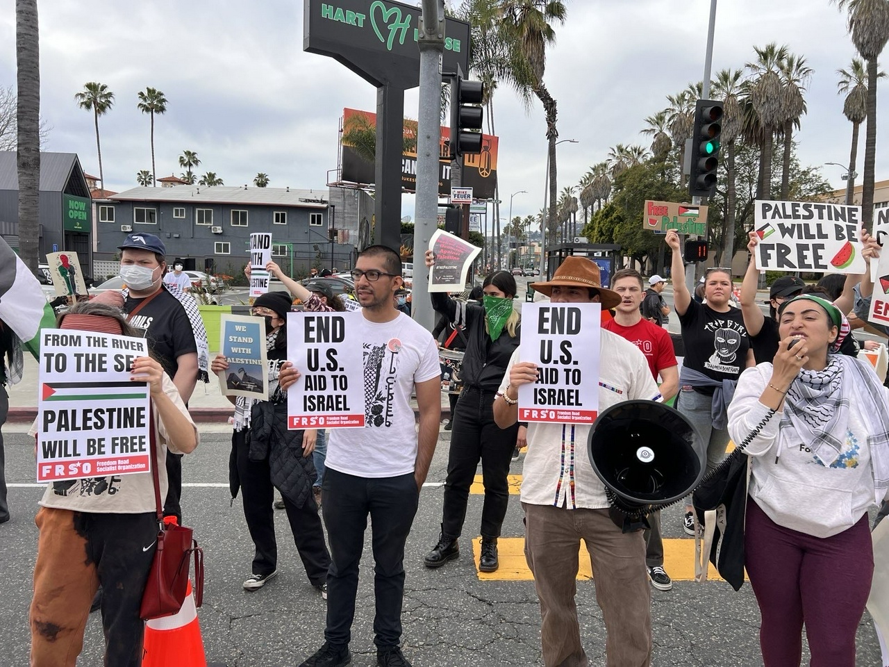 Protesters hold signs saying "From the river to the sea Palestine will be free" and "End US Aid to Israel" while a lead protester chants on a bullhorn.