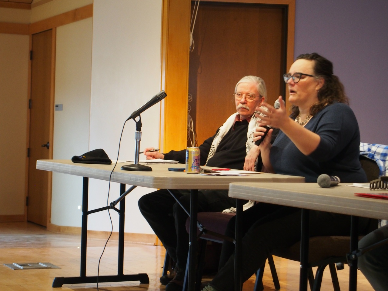 Two people sitting at a table, with one speaking into a microphone. 