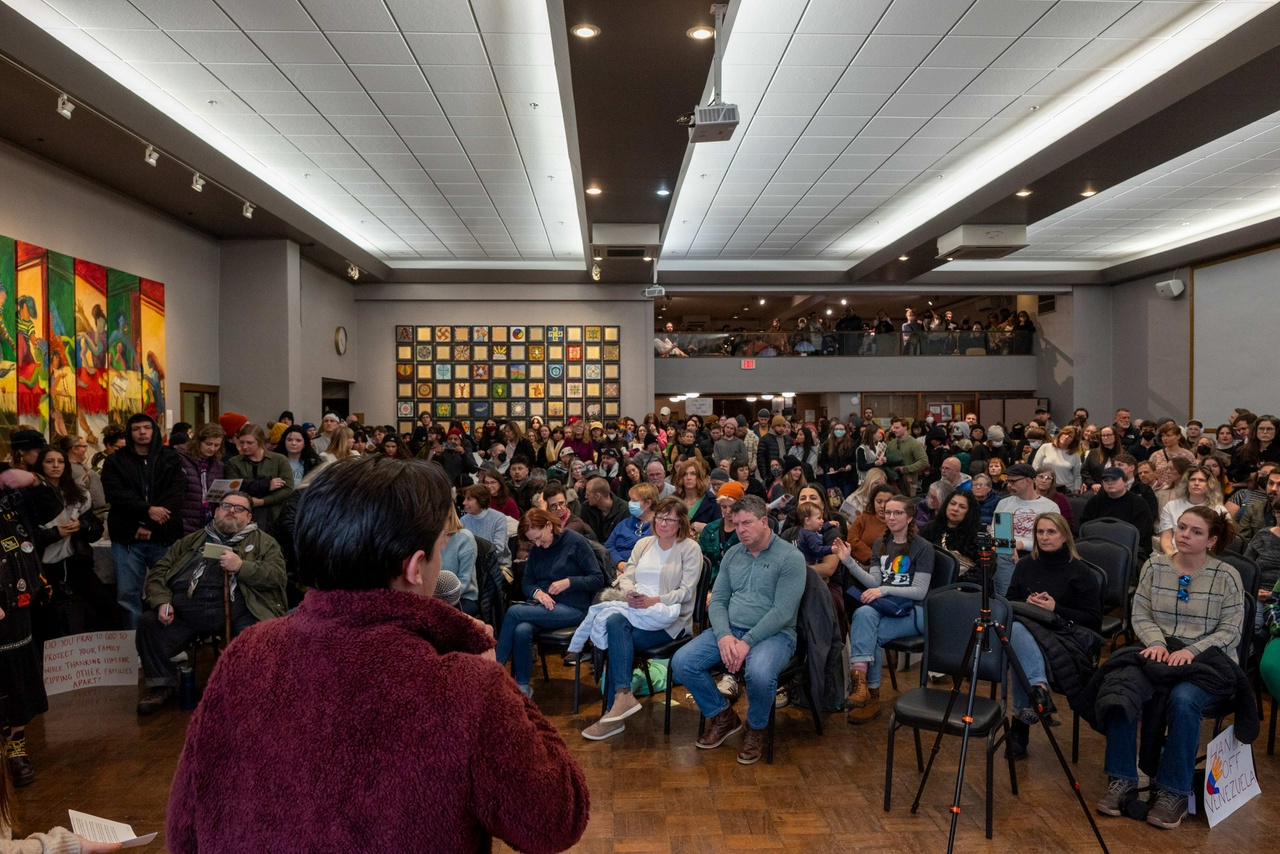 Person speaks on a mic at the front of a crowded room. The people in the room sit attentively.