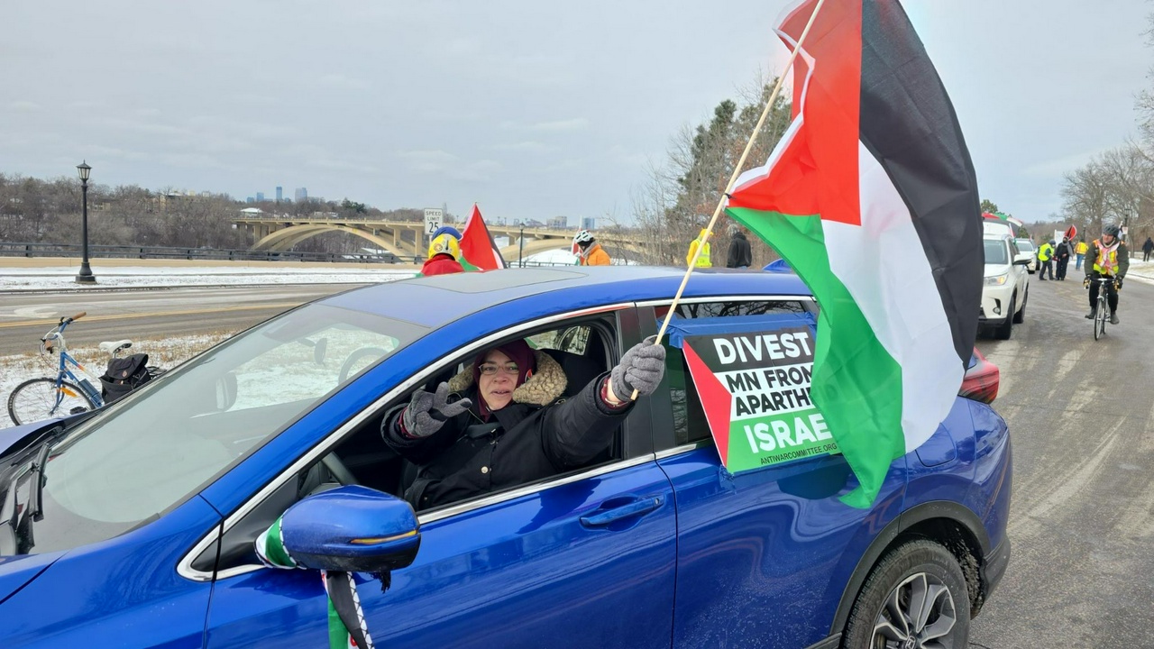 Woman wearing a red hijab waves a Palestinian flag out of the driver window of the blue car she is driving.