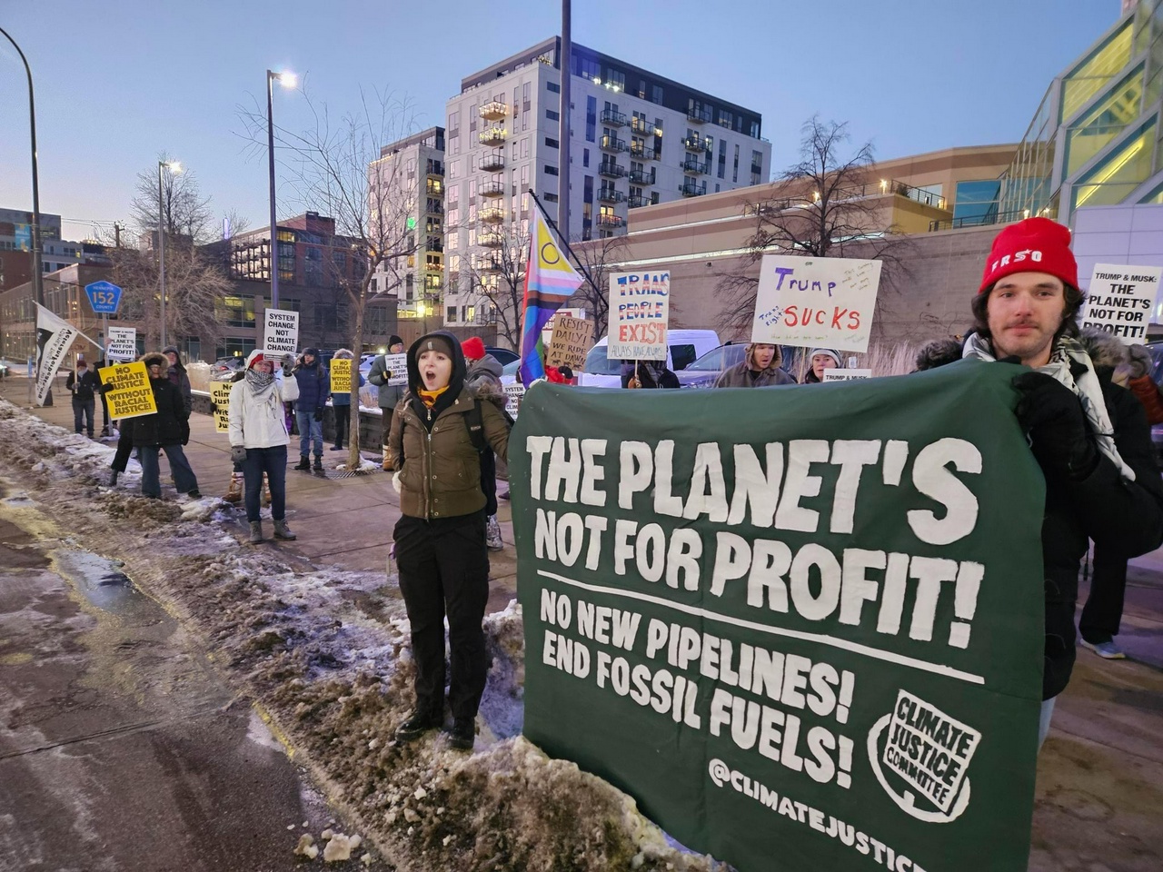 Protesters line up along a roadside carrying signs and banners reading things like “The Planet’s Not For Profit! No New Pipelines! End Fossil Fuels”.