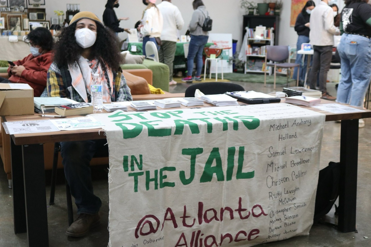 Atlanta Alliance organizer wears a mask and sits behind a table with a banner on it’s front that reads “Stop the deaths in the jail @atlantaalliance”
