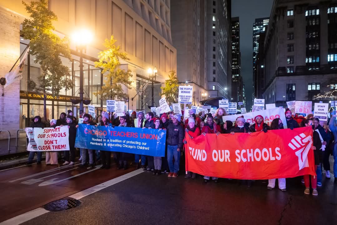 Crowd marches in street behing banners that read “Fund Our Schools” and “Chicago Teachers Union - rooted in a tration of protecting out members and educating Chicago’s Children” and “Support our schools - don’t close them”.