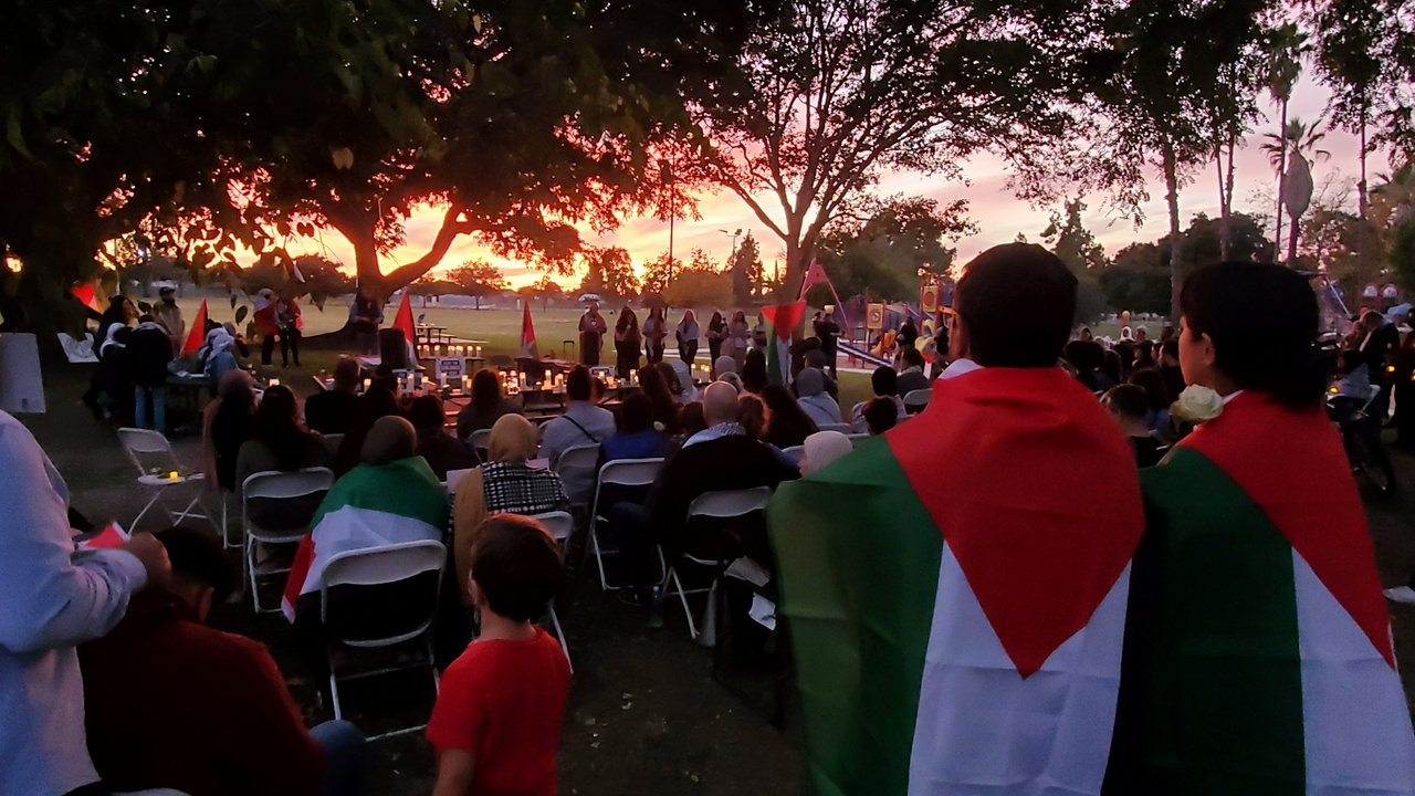 Crowd wearing Palestinian flags, seated and standing, listen as a speaker talks up front.