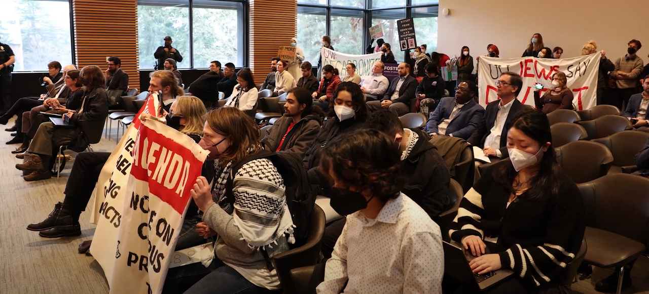 A crowd of people sits in chairs. People hold large banners in the front and back rows.