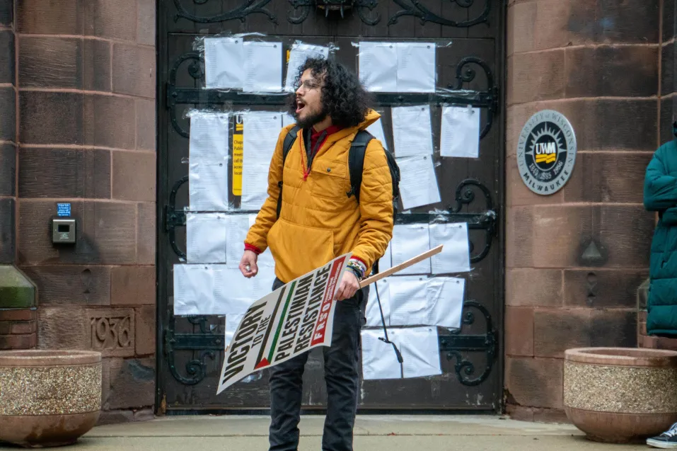 Student holds sign that reads “Victory to the Palestinian Resistance” while standing in front of a UWM building door covered in petition signatures.