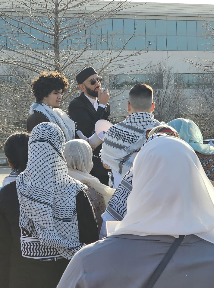 A group of youth in khaffiyehs gather and listen to speakers.