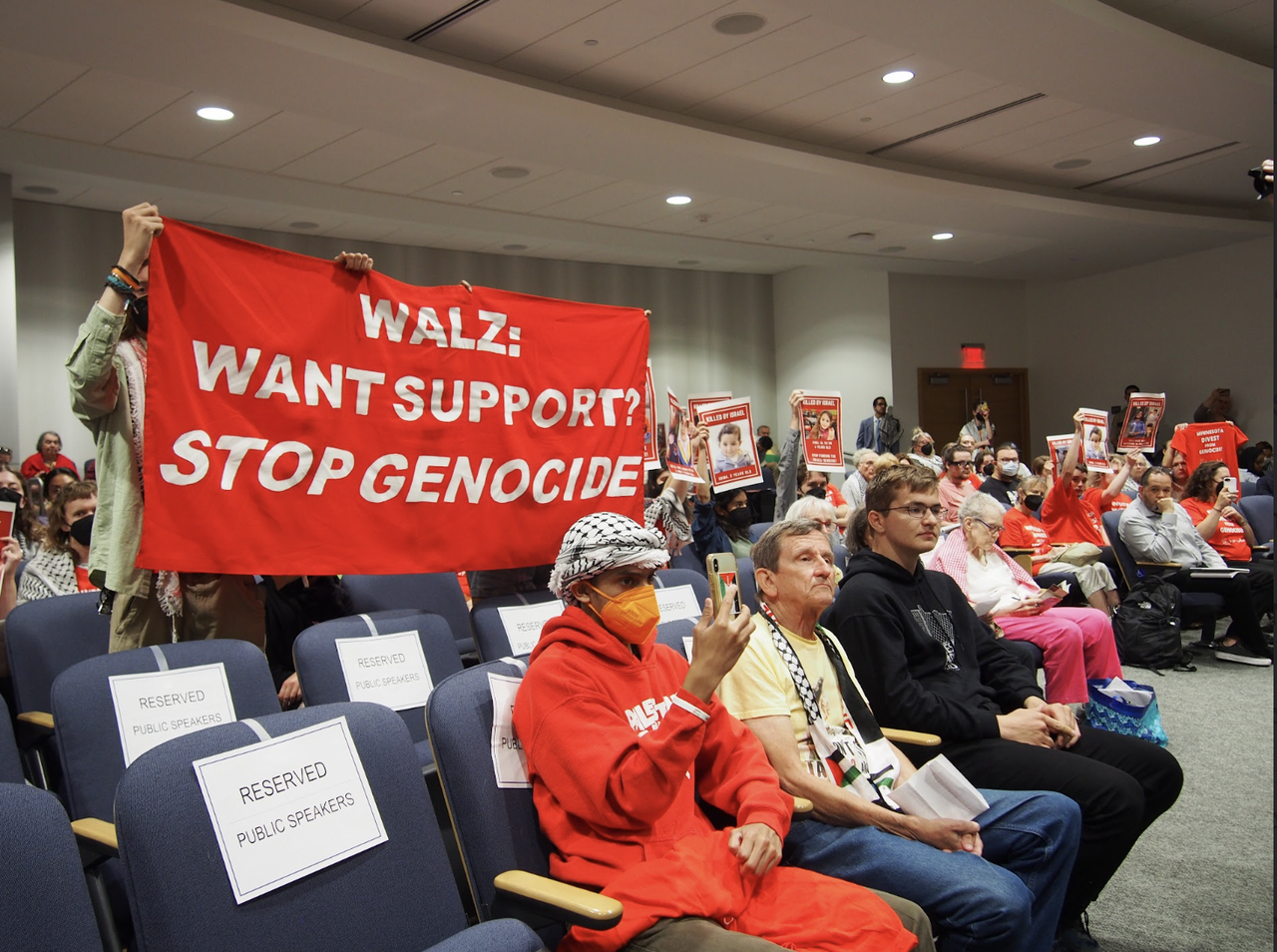 Audience sits at meeting while protestors in audience hold up banner that reads "Walz: Want support? Stop genocide."