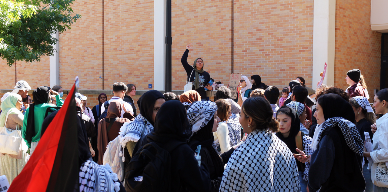 Students wearing keffiyehs and waving Palestinian flags surround one student giving a speech in front of a brick building