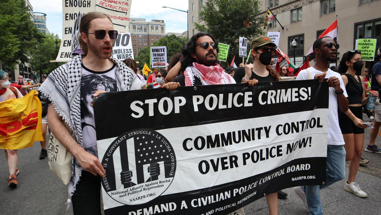 Protestors carry a banner that says “Stop Police Crimes! Community control over police now!” Dozens of other protestors are visible marching behind and beside them.